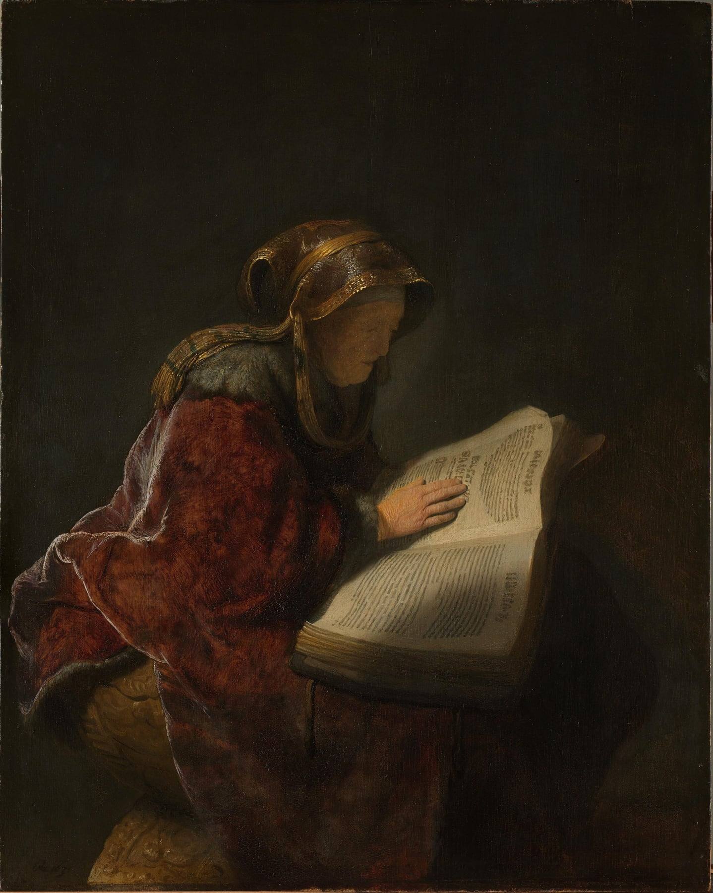 Rembrandt’s painting of an elderly woman in a red fur-trimmed robe and gold-embroidered headscarf, seated in profile in a dark room, one hand resting on the brightly lit pages of a large open book.